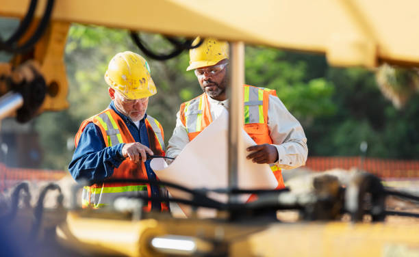 Two multiracial construction workers, in their 50s, looking at blue prints at a construction site. The African-American man is holding the plans open while his Hispanic coworker looks and points.